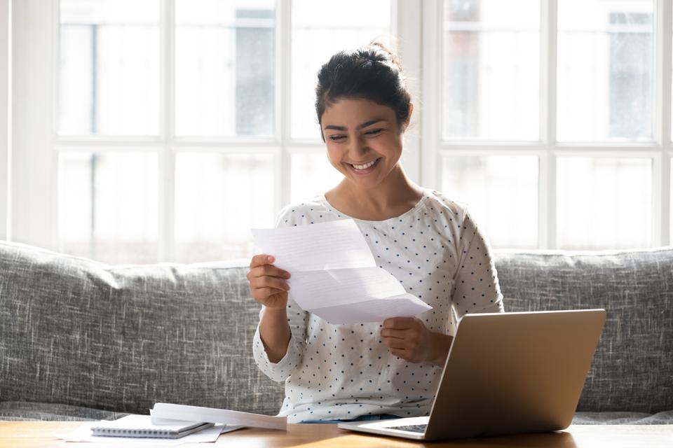 Smiling woman reading letter near laptop in sunny room